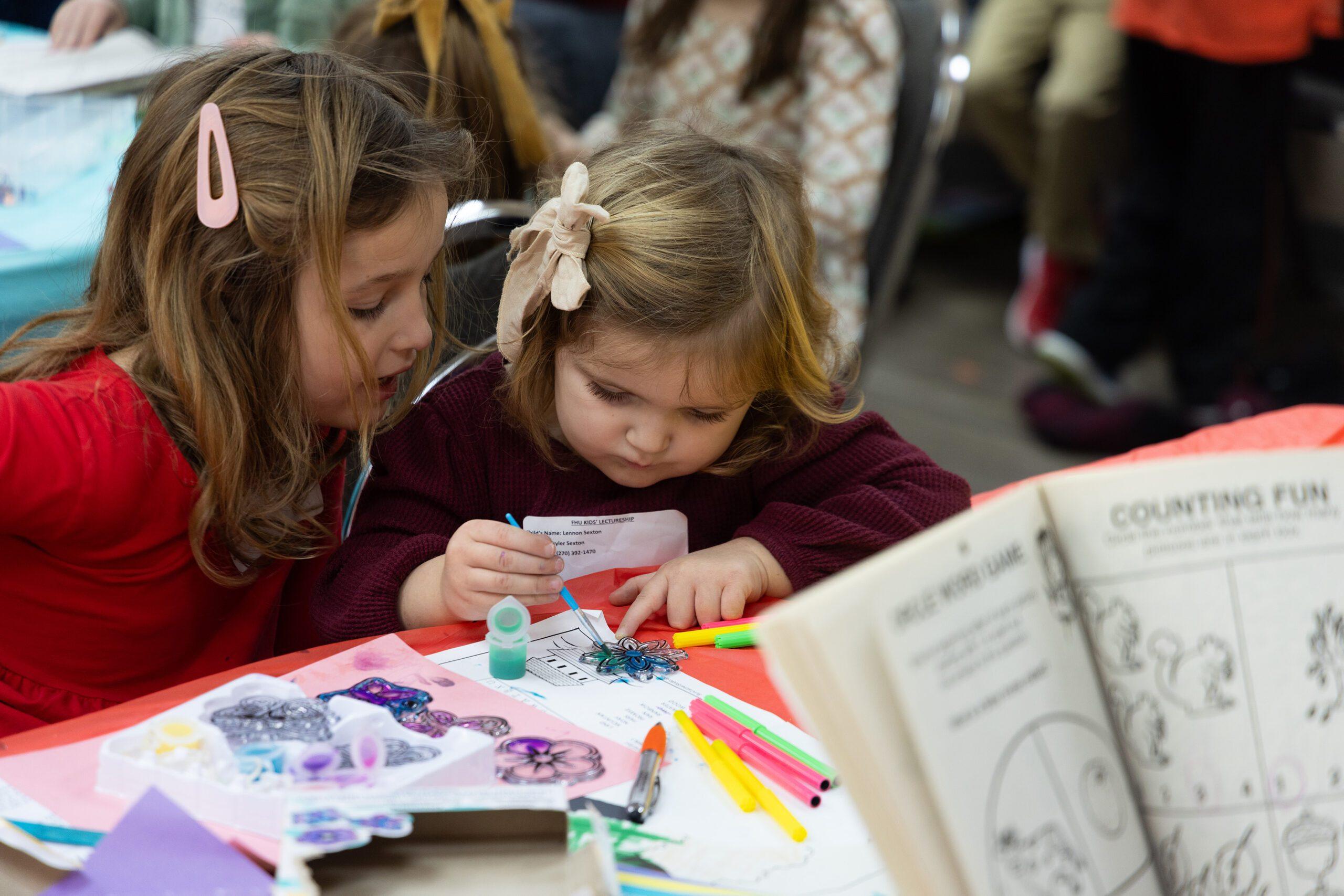 Two young girls sit at a table covered with art supplies, focusing intently on coloring and drawing together. One girl watches closely while the other uses a blue marker on a paper. An open activity book is visible in the foreground.