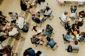 A group of people sit in pairs on chairs arranged in rows in a large indoor space, engaging in face-to-face conversations, with papers and notebooks in hand. The scene is viewed from above.