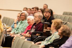 A group of older adults, including a man in a wheelchair, sit in an auditorium among other attendees, all facing forward and appearing engaged in the event.