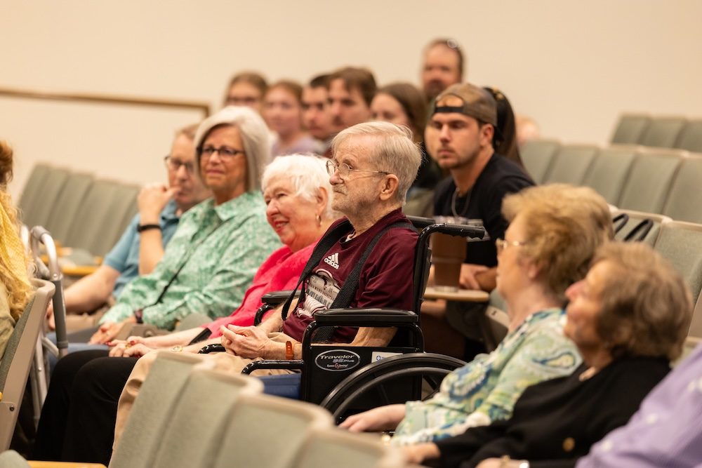 A group of older adults, including a man in a wheelchair, sit in an auditorium among other attendees, all facing forward and appearing engaged in the event.