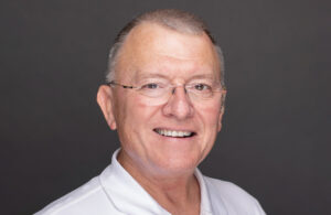An older man with short gray hair, glasses, and a white collared shirt smiles warmly against a plain dark background.