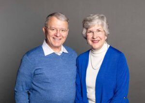 An older man and woman stand side by side, smiling at the camera. Both have gray hair and wear glasses. The man wears a blue sweater over a collared shirt; the woman wears a blue cardigan over a white turtleneck.