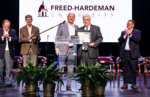 Two men stand center stage at a Freed-Hardeman University event, one handing the other a framed certificate, while three other men beside them applaud. Potted plants are on the stage in front of them.