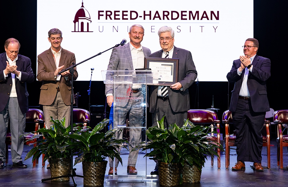 Two men stand center stage at a Freed-Hardeman University event, one handing the other a framed certificate, while three other men beside them applaud. Potted plants are on the stage in front of them.
