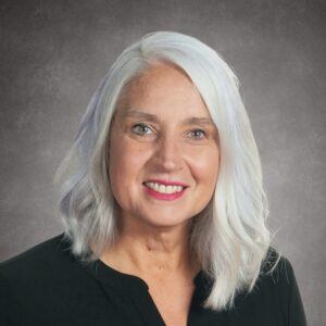 A woman with shoulder-length white hair and a bright smile is wearing a black top, posing against a gray studio background.