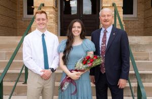 Three people stand on steps outside a brick building. The woman in the center holds a bouquet of red roses and wears a blue dress. The two men on either side wear a suit and a shirt with a tie. All three are smiling at the camera.