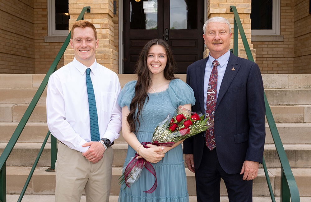 Three people stand on steps outside a brick building. The woman in the center holds a bouquet of red roses and wears a blue dress. The two men on either side wear a suit and a shirt with a tie. All three are smiling at the camera.