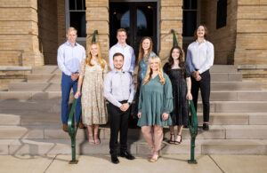 Eight young adults, four men and four women, stand and sit on the steps of a brick building, posing for a group photo. They are dressed in semi-formal attire and smiling at the camera.