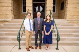 Three people stand in front of stone steps and a brick building. The man in the center wears a suit, the young man on the left wears a white shirt and khakis, and the woman on the right wears a blue dress.