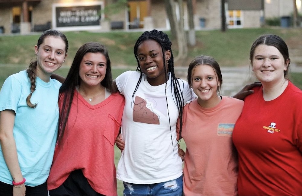 Five young women stand close together outdoors, smiling at the camera. They are casually dressed in t-shirts, and greenery and a building are visible in the blurred background.