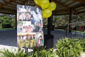 A photo collage on an easel stands outdoors under a pavilion, surrounded by yellow balloons and ferns. The collage features various group and family photos. The area is sunny with trees in the background.