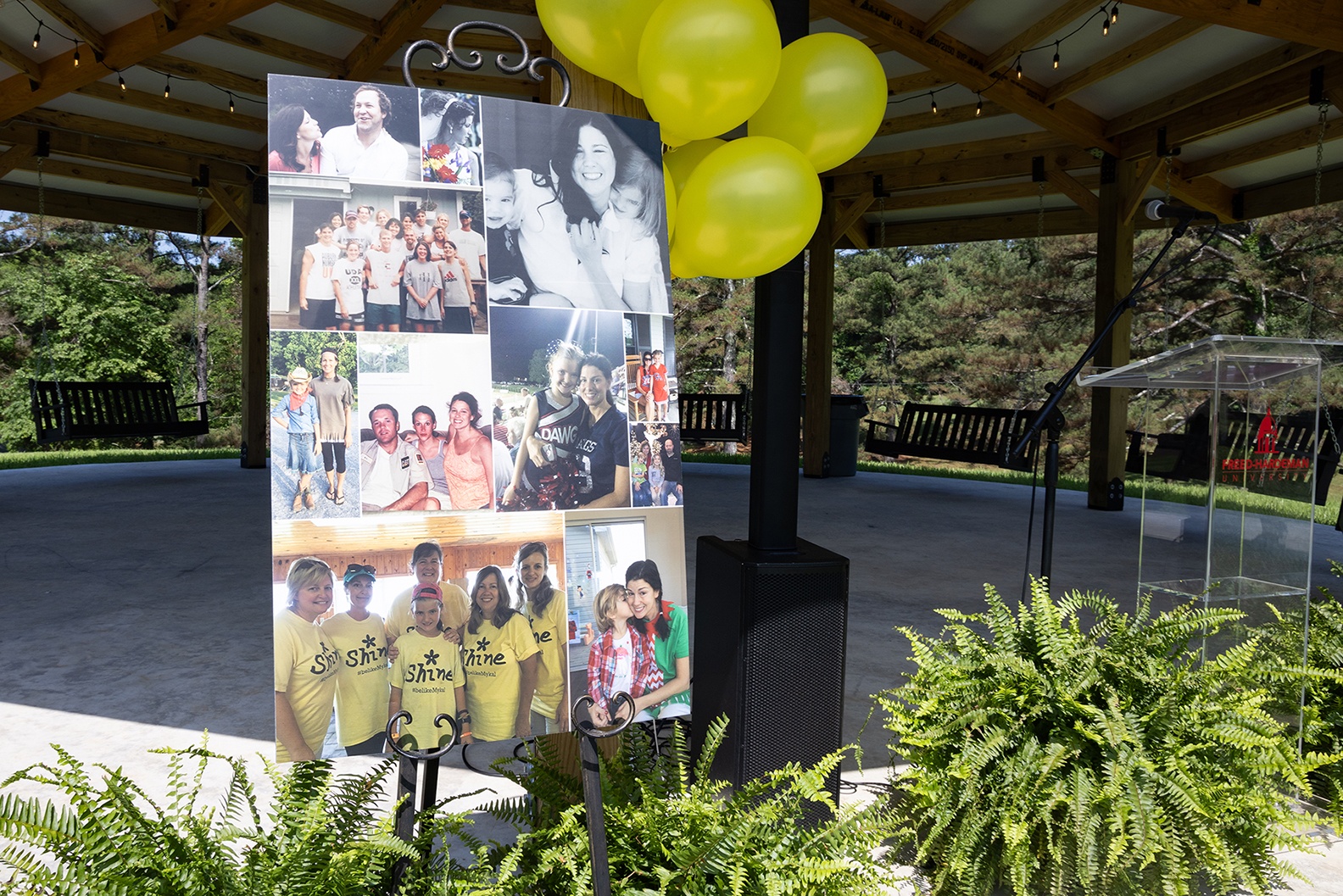 A photo collage on an easel stands outdoors under a pavilion, surrounded by yellow balloons and ferns. The collage features various group and family photos. The area is sunny with trees in the background.
