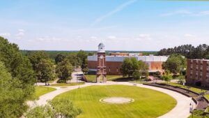 Aerial view of a college campus with a large circular grassy quad, a brick building with a clock tower in the center, and surrounding trees and academic buildings under a clear blue sky.