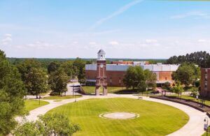 Aerial view of a university campus featuring a large grassy circle, a brick clock tower with a white dome, brick buildings, trees, and a clear blue sky.