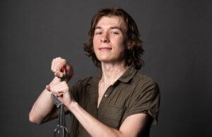 A young person with medium-length brown hair and a brown short-sleeve shirt holds a kitchen whisk and smiles slightly in front of a plain dark background.