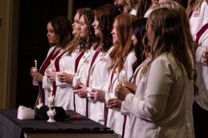 A group of women in white coats stand in a row, each holding a lit candle during a ceremony. They are facing forward, appearing attentive and focused, with maroon sashes over their shoulders.
