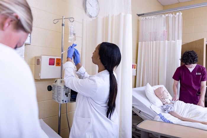 A healthcare worker adjusts an IV bag while another person stands by a hospital bed with a medical mannequin, suggesting a medical training or simulation setting. Curtains and medical equipment are visible in the room.