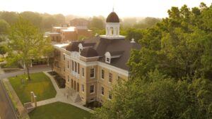 Aerial view of a historic brick building with a white cupola and black roof, surrounded by lush green trees and bathed in warm sunlight. Stairs lead to the main entrance, and sidewalks border the landscaped grounds.