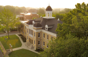 Aerial view of a historic brick building with a domed cupola, surrounded by trees and green lawns, bathed in warm sunlight, with modern buildings visible in the background.