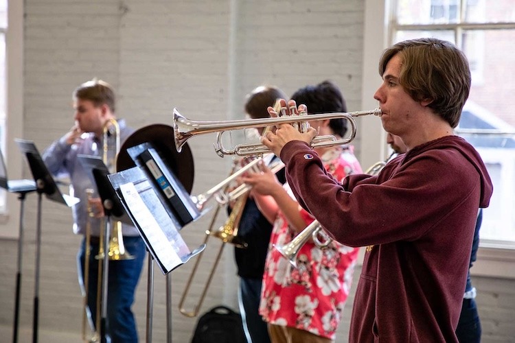 A group of musicians practice indoors, playing brass instruments including trumpets and trombones. Music stands with sheet music are in front of them, and natural light comes through nearby windows.