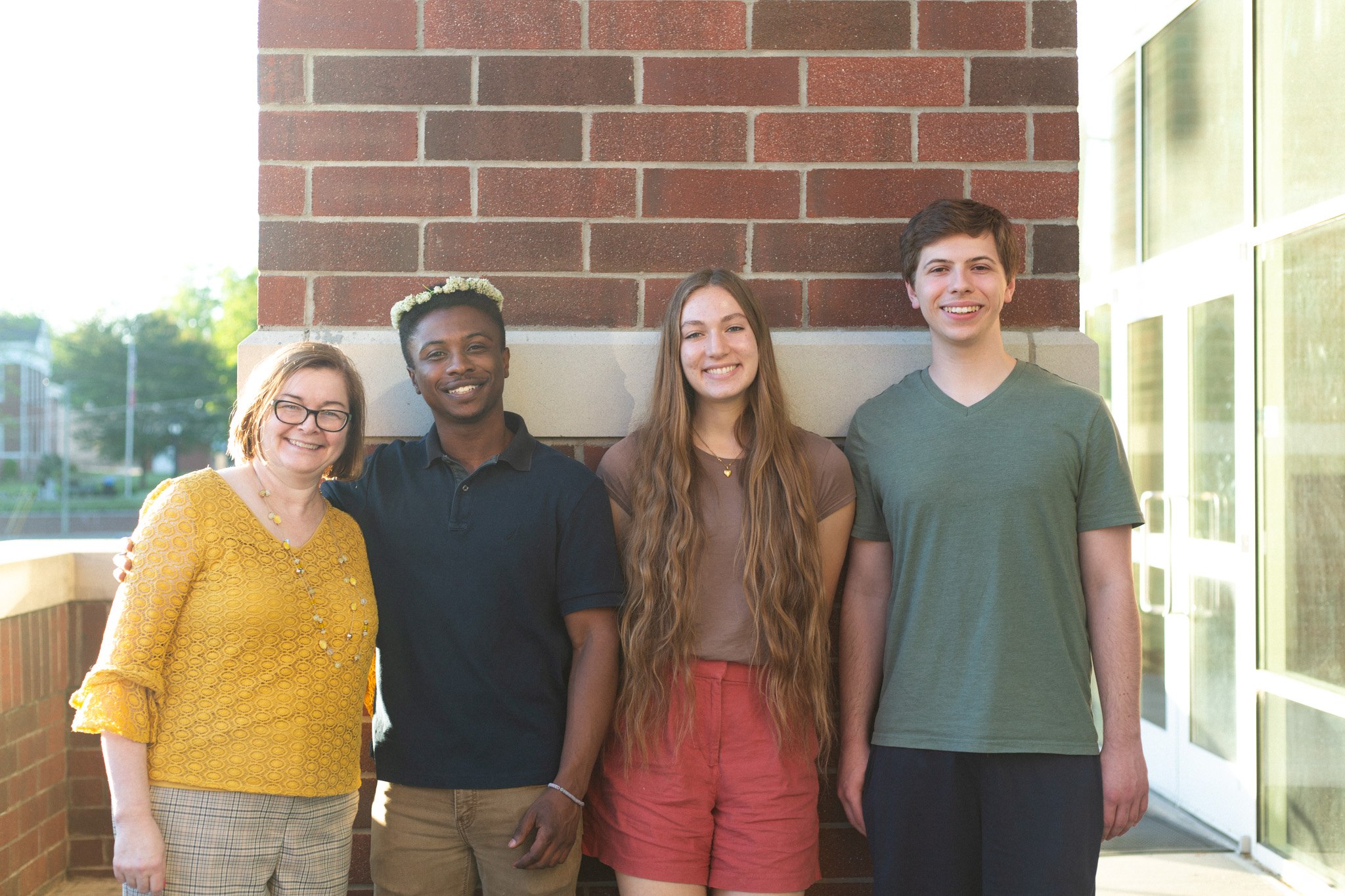 Four young adults stand side by side, smiling at the camera in front of a brick wall on a sunny day. They appear relaxed and friendly, dressed in casual summer clothes.
