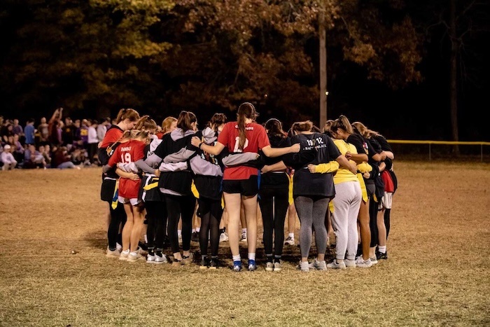 A group of young women in sports uniforms stand in a circle on a field at night, arms around each other’s shoulders, with trees and spectators visible in the background.
