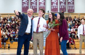 A man takes a selfie with three young people in a gymnasium. One young woman holds flowers. Bleachers full of people and banners are visible in the background. Everyone is smiling and dressed up for the occasion.