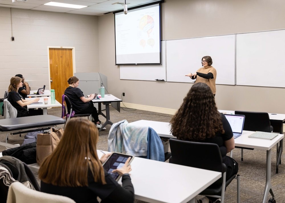 A teacher stands at the front of a classroom, gesturing as she speaks to seated students who are taking notes on laptops and tablets. A brain diagram is projected onto the whiteboard behind her.