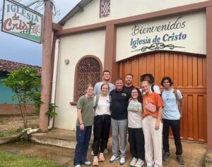 A group of nine smiling people stand together in front of a building with a sign reading Bienvenidos Iglesia de Cristo above the door. The building has a white and brown exterior.