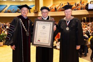 Three men in academic regalia stand on a stage at a graduation ceremony. The man in the center holds a large framed diploma. An audience is seated in the background.