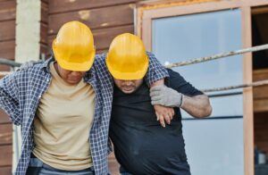 Two construction workers wearing yellow hard hats support each other, with one worker appearing injured or tired. They are standing close together in front of a wooden building.
