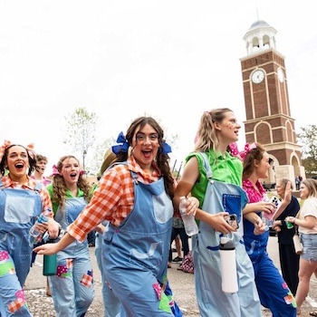 A group of young women in colorful overalls and shirts, some with bows in their hair, walk and smile outdoors near a tall brick clock tower, appearing festive and energetic.