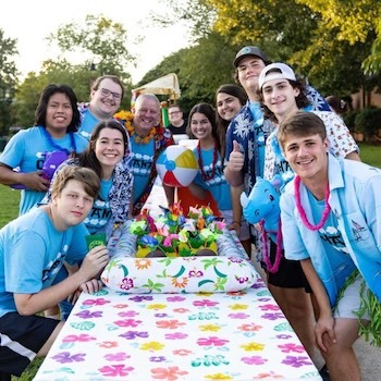 A group of smiling young people in blue shirts and leis gather around a colorful, flower-patterned table outdoors, with a beach ball and summer decorations, posing for a cheerful group photo.