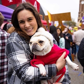 A smiling woman in a plaid coat holds a French bulldog wearing a red sweater and knit hat at an outdoor event with crowds and colorful tents in the background.