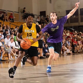 Two men play basketball in a gym. The player in a yellow Sigma Rho jersey dribbles the ball, while the player in a purple shirt defends. Spectators fill the bleachers in the background.