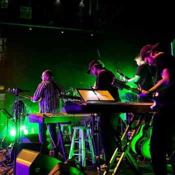 A band performs on stage with green lighting. Four musicians play guitars and keyboards, and various musical equipment is visible. The background is dark, emphasizing the vibrant stage lights.