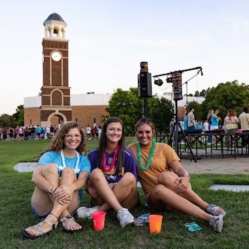 Three young women sit on grass smiling at the camera, with colorful cups in front of them. A crowd and a brick clock tower are visible in the background under a clear sky.