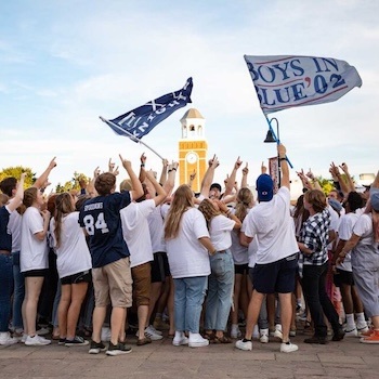 A large group of people in casual clothes gather outdoors, raising their arms and waving flags. A clock tower stands in the background under a blue sky.