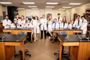 A group of students and instructors in white lab coats pose together in a science laboratory classroom, standing and smiling behind lab benches.