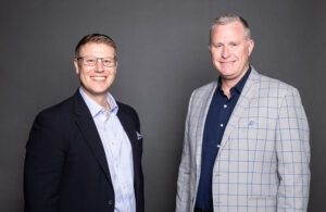 Two men stand side by side against a plain gray background. Both are smiling and wearing jackets; the man on the left has glasses and a dark blazer, while the man on the right wears a light plaid blazer.