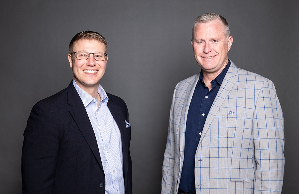 Two men stand side by side against a plain gray background. Both are smiling and wearing jackets; the man on the left has glasses and a dark blazer, while the man on the right wears a light plaid blazer.