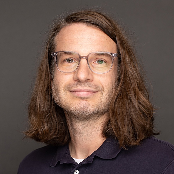 A man with shoulder-length brown hair, wearing clear-framed glasses and a dark polo shirt, smiles slightly against a plain gray background.