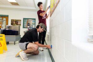 Two people are cleaning a white brick wall in a bright room. One is standing and wiping near a bulletin board, while the other is crouching and cleaning lower on the wall. A yellow caution sign is visible on the floor nearby.