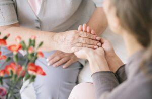Close-up of a younger person gently holding the hands of an older person, offering comfort and support. Pink flowers are visible in the foreground.