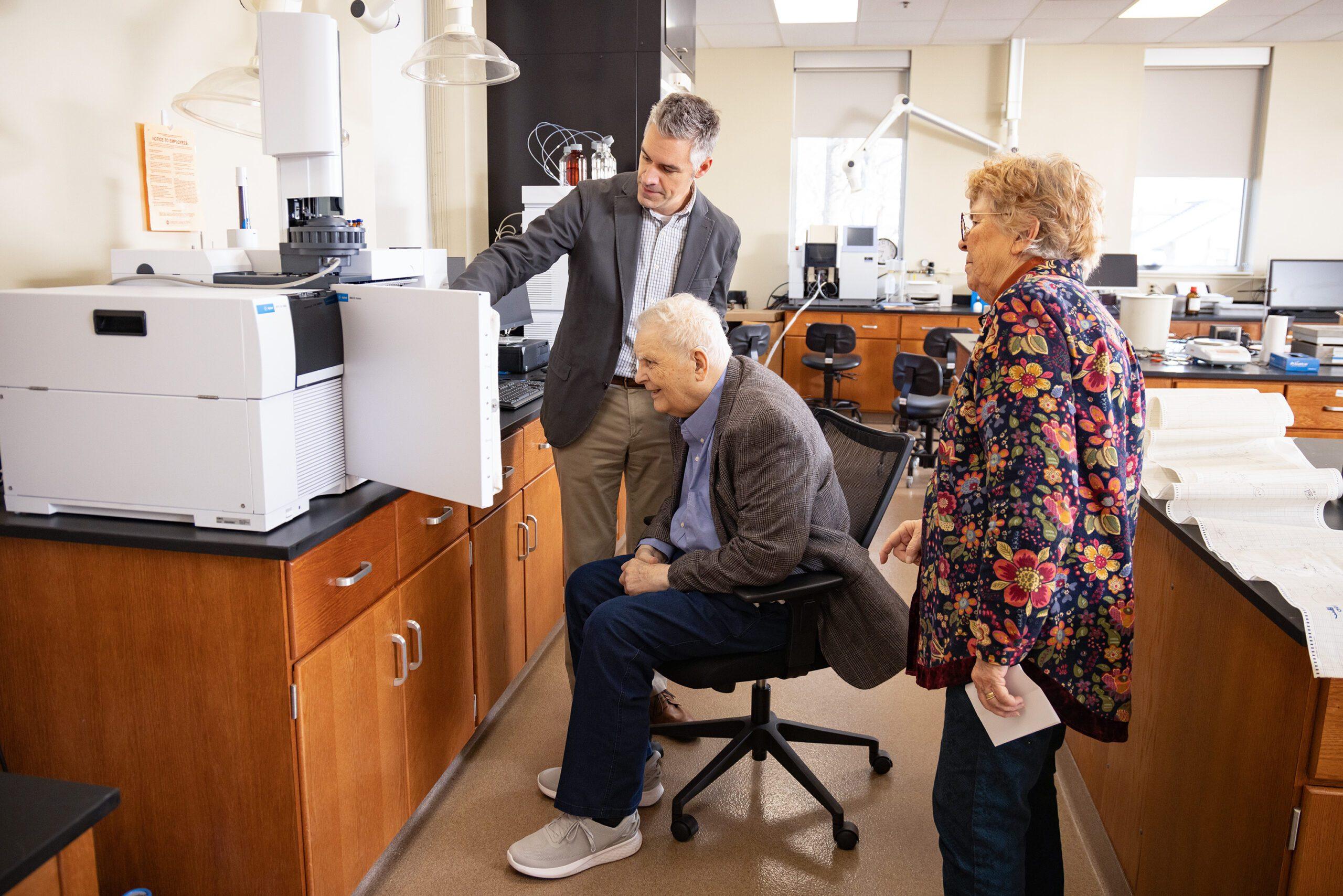 Three people in a laboratory observe a scientific instrument; one man stands pointing at the machine, another man sits in a chair, and a woman stands nearby, all focused on the equipment.