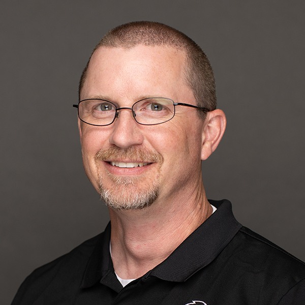 A man with short brown hair, glasses, and a trimmed goatee smiles while wearing a black collared shirt against a plain gray background.