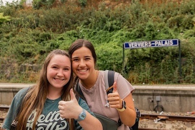 Two young women standing on a train platform, smiling and giving thumbs up. A sign behind them reads Verviers-Palais. Greenery covers the slope in the background.