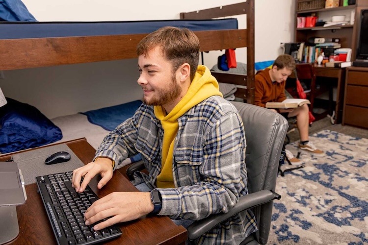 A young man in a yellow hoodie and plaid shirt types on a keyboard at a desk in a dorm room. In the background, another student sits on a chair reading a book next to a bunk bed.