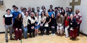 A group of about thirty young adults and a couple of adults pose and smile together indoors against a gymnasium wall. Some are standing, while others are kneeling in front.