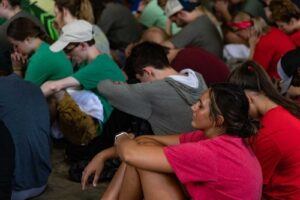 A group of young people sit closely together on the ground, many with heads bowed and eyes closed, appearing thoughtful or in prayer. Most are dressed casually in colorful t-shirts.